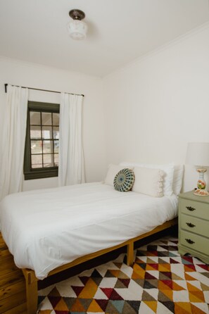 Une chambre à coucher avec un lit, une fenêtre avec des rideaux, une table de chevet avec une lampe et un sol à motifs.