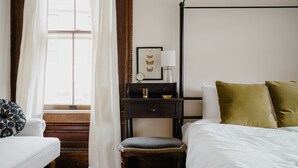 Ein Schlafzimmer mit einem Bett, einem Sessel, einem Nachttisch, einem Fenster mit Vorhängen und einer Wandleuchte.