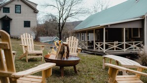 Ein Feuerkorb mit Holzscheiten und einer Metallschale, umgeben von Holz-Adirondack-Stühlen auf einer grünen Wiese, mit einem Haus im Hintergrund.