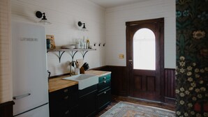 Eine Küche mit einem weißen Kühlschrank, einer Spüle und Holzschränken. Eine Tür mit dekorativer Verkleidung und ein Teppich liegen auf dem Boden.