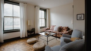 Ein Wohnzimmer mit Sofa, Hocker und Couchtisch. Es gibt zwei Fenster mit Vorhängen, eine Lampe und ein gerahmtes Bild an der Wand.