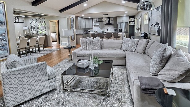 A spacious living area with a large L-shaped sofa, a coffee table, and a dining table with chairs. The room features a high ceiling with exposed beams, recessed lighting, and a view of the kitchen area.