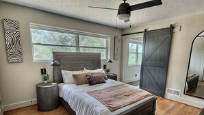 A bedroom with a large bed, a ceiling fan, a sliding barn-style door, and a large window with a view of trees.