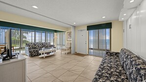 A spacious living area with a sofa, a coffee table, and a chair. There is a large window with blinds, a door, and a view of the ocean. - Studio by the Sea | Beach, Pool & Sunset Vibes (Longboat Key)