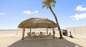 A beach umbrella with chairs and a grill on a sandy beach.