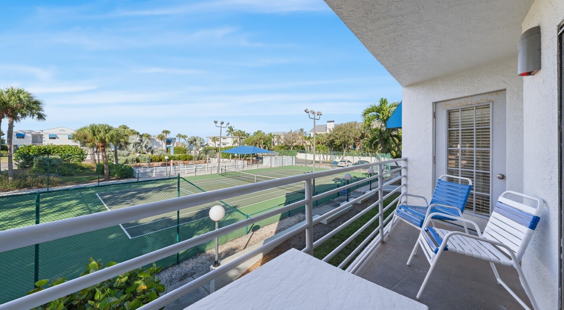 A balcony with a tennis court view, outdoor seating, and a clear sky.