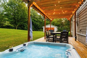 A hot tub with a wooden deck, a covered seating area with chairs, and a house in the background.