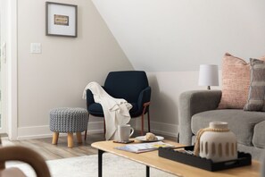 A modern living room with a dark blue armchair, a grey sofa, a small ottoman, and a coffee table with a mug and a plate of food.