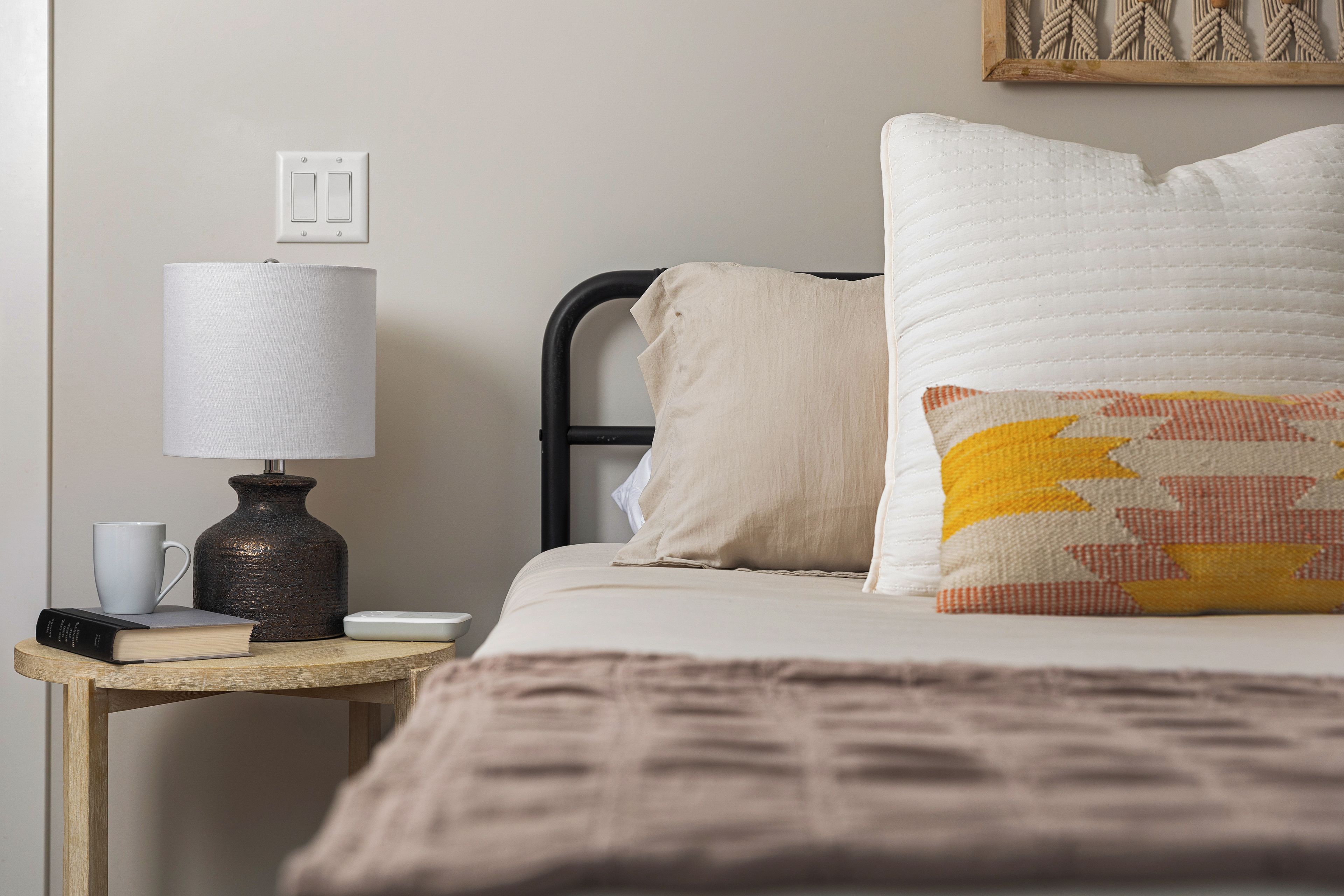 Un lit bien fait, recouvert d’une couverture beige, avec un oreiller à motifs géométriques et un autre oreiller uni. Une table de chevet porte une lampe, une tasse et un livre.