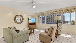 A living room with a ceiling fan, a television, a sofa, a chair, and a window with a view of a building and trees. - Welcome to your happy place—aka where real life takes a vacation. (Sarasota)