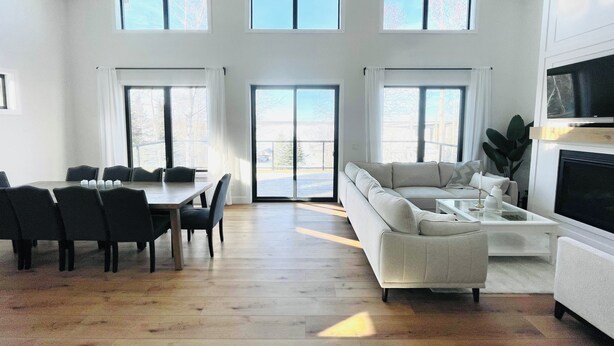A spacious living area with a high ceiling, large windows, a dining table with chairs, a sofa, and a television.