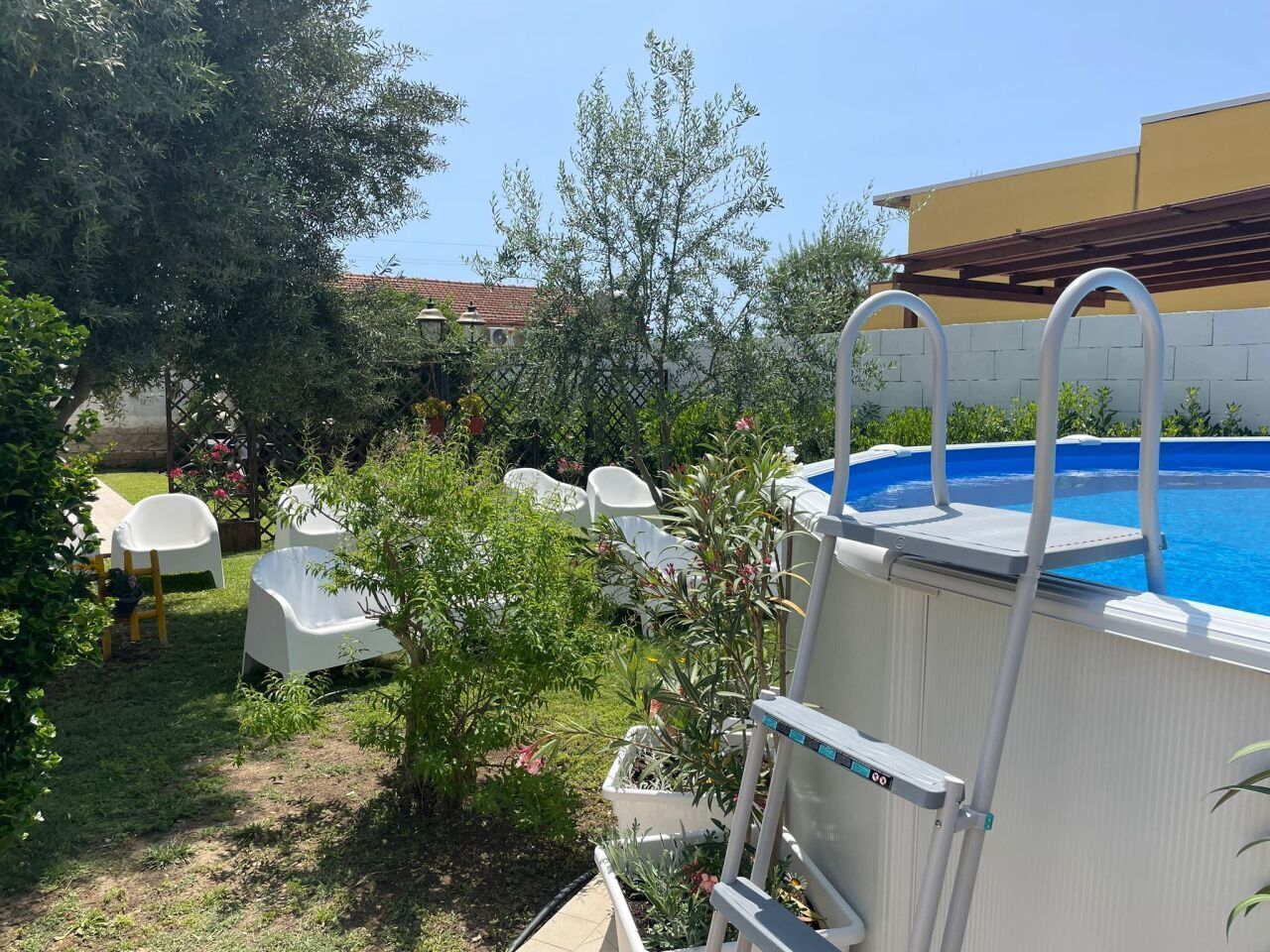 An outdoor swimming pool with a ladder, surrounded by greenery and white plastic chairs.