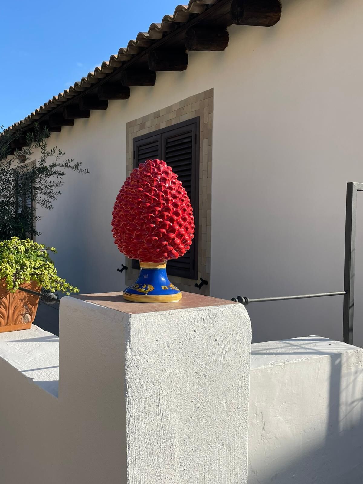 Ein Betonblock mit einer roten Kugel darauf, einem Blumentopf und einem Gebäude mit Ziegeldach und Fensterläden.