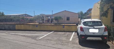 A parking lot with a white car parked near a building with a yellow wall and a fence.