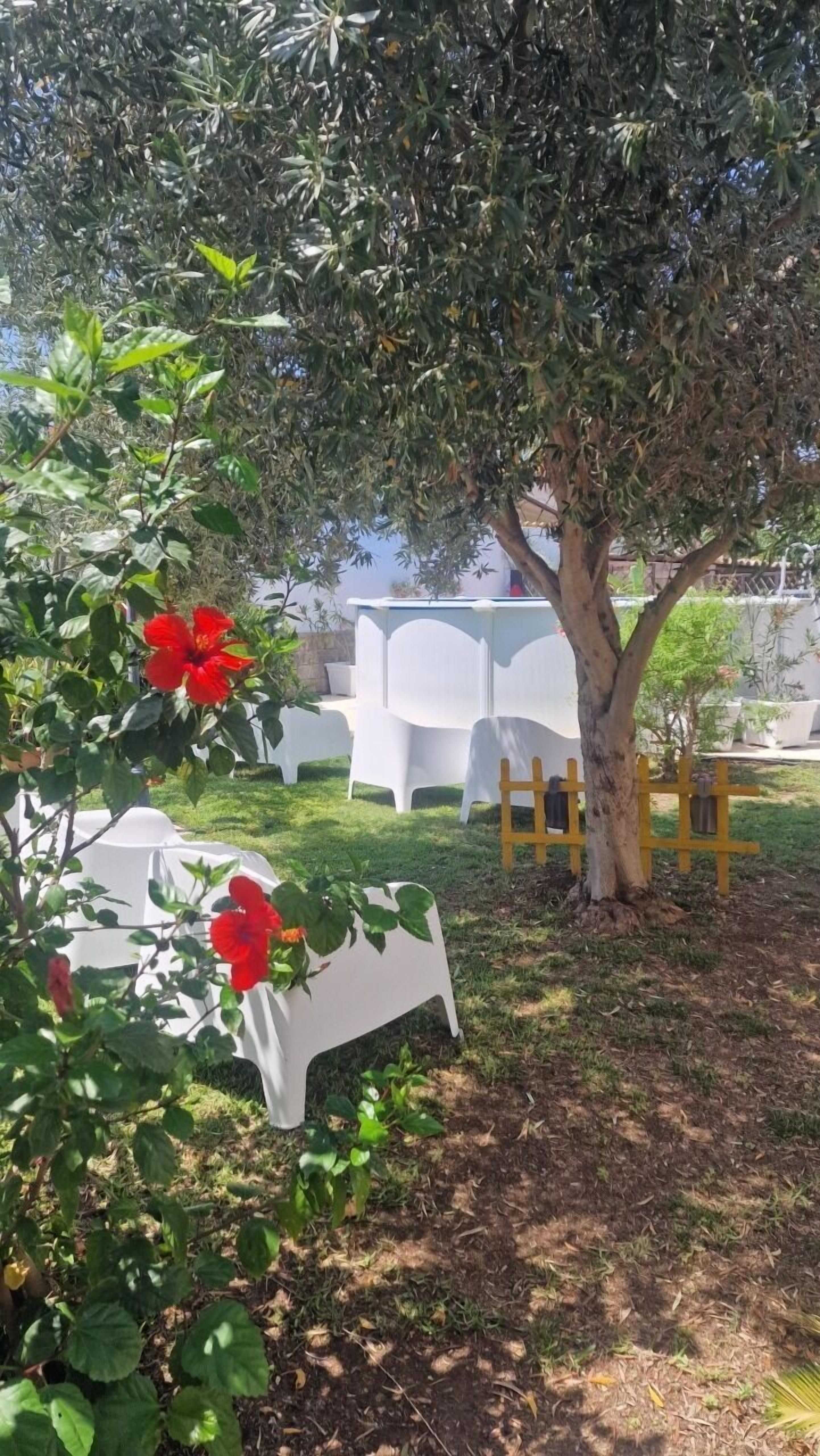 Un espace extérieur aménagé avec des chaises et des tables en plastique blanc, entouré de verdure et d’un arbre.