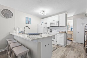 A modern kitchen with white cabinetry, a central island with stools, a microwave, and a clock on the wall. - Incredible Ocean Front Views, Indoor/Outdoor Pool, Hot Tub, Fully Stocked, 4 TVs (Orange Beach)