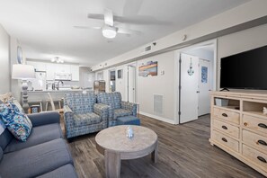 A modern living room with a sofa, armchair, and coffee table. A TV is mounted on the wall, and there is a kitchen in the background.
