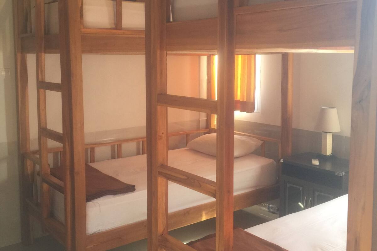 Une chambre avec trois lits superposés en bois, une table de chevet avec une lampe et une petite commode.