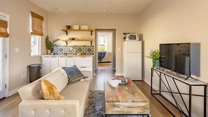 Ein modernes Wohnzimmer mit Sofa, Couchtisch und Fernseher. Im Hintergrund ist eine Küche mit weißen Schränken und einer Mikrowelle zu sehen.