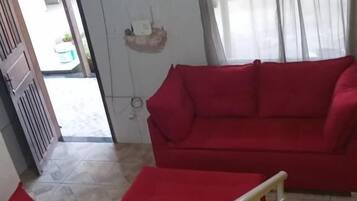 Una sala de estar con un sofá rojo, un sillón amarillo y un cojín floral, y suelo de baldosas.
