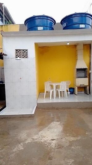 Una pequeña área de cocina al aire libre con una pared amarilla, una pared de azulejos blancos, una mesa con sillas y una parrilla.