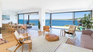 A spacious living area with a large window overlooking a body of water, wooden furniture, and a cozy seating arrangement. - 6 bedrooms, ocean views, pool & spa (Cronulla)