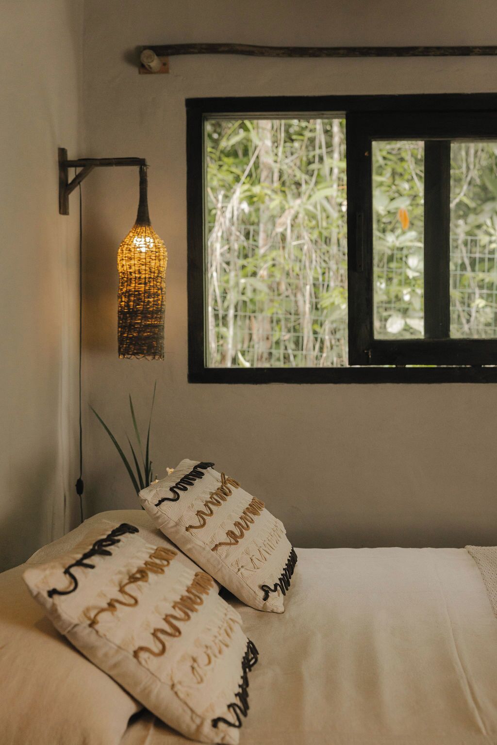 Un dormitorio con cama, dos almohadas con texto bordado, una lámpara colgante y una ventana con vista a follaje.