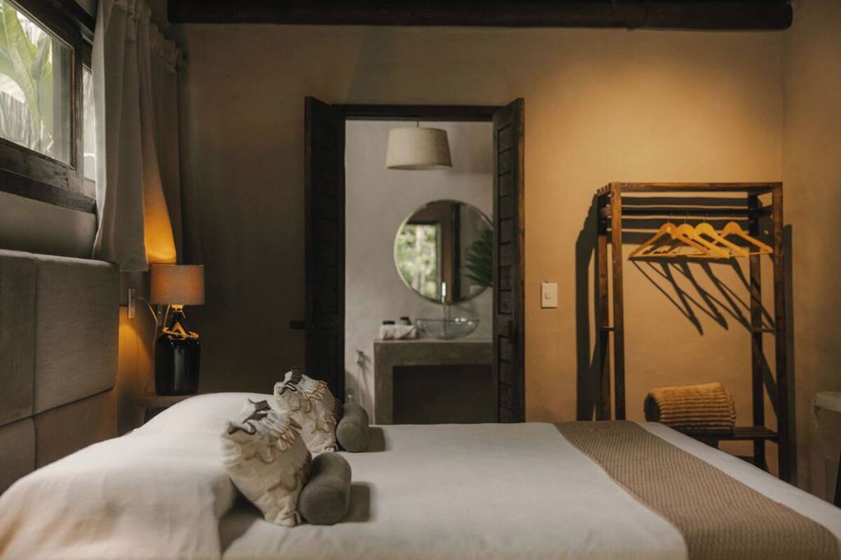 A neatly made bed with pillows, a bedside lamp, a mirror, and a wooden rack with hangers.
