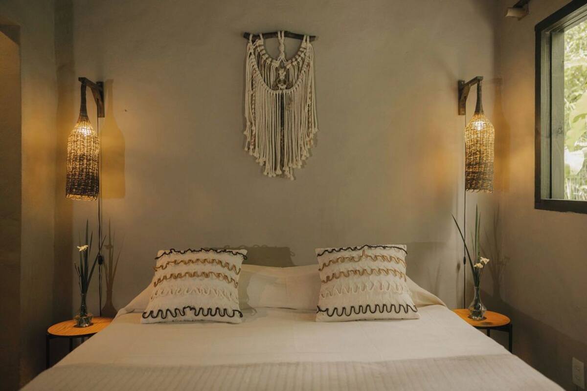 A bedroom with a bed, two bedside lamps, a macrame wall hanging, and a window.