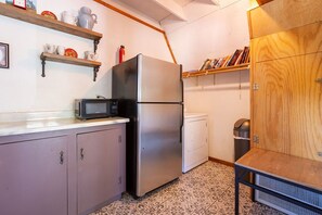 Una cocina pequeña y funcional con refrigerador, microondas y lavadora. Se observan estantes de madera con libros y tazas, además de un piso con diseño.