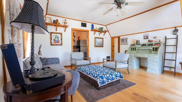 A room with a ceiling fan, a bed with a patterned bedspread, a desk with books and a lamp, and a chair. - The Library - A Hill Country Cottage (San Marcos)
