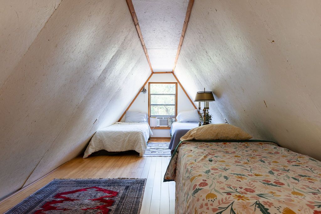 Un dormitorio pequeño en el ático, con forma de triángulo, que tiene dos camas, una ventana y una lámpara.