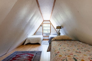 A small, triangular attic bedroom with two beds, a window, and a lamp. - The Library - A Hill Country Cottage (San Marcos)