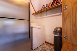 A compact laundry room with a washing machine, a trash bin, and a wooden shelf above the washing machine.