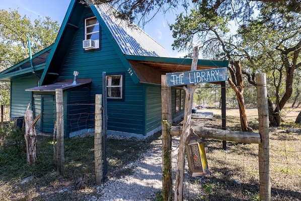 Un bâtiment rustique, sur lequel on peut lire l’inscription « La bibliothèque », entouré d’arbres et bordé d’un chemin de gravier.