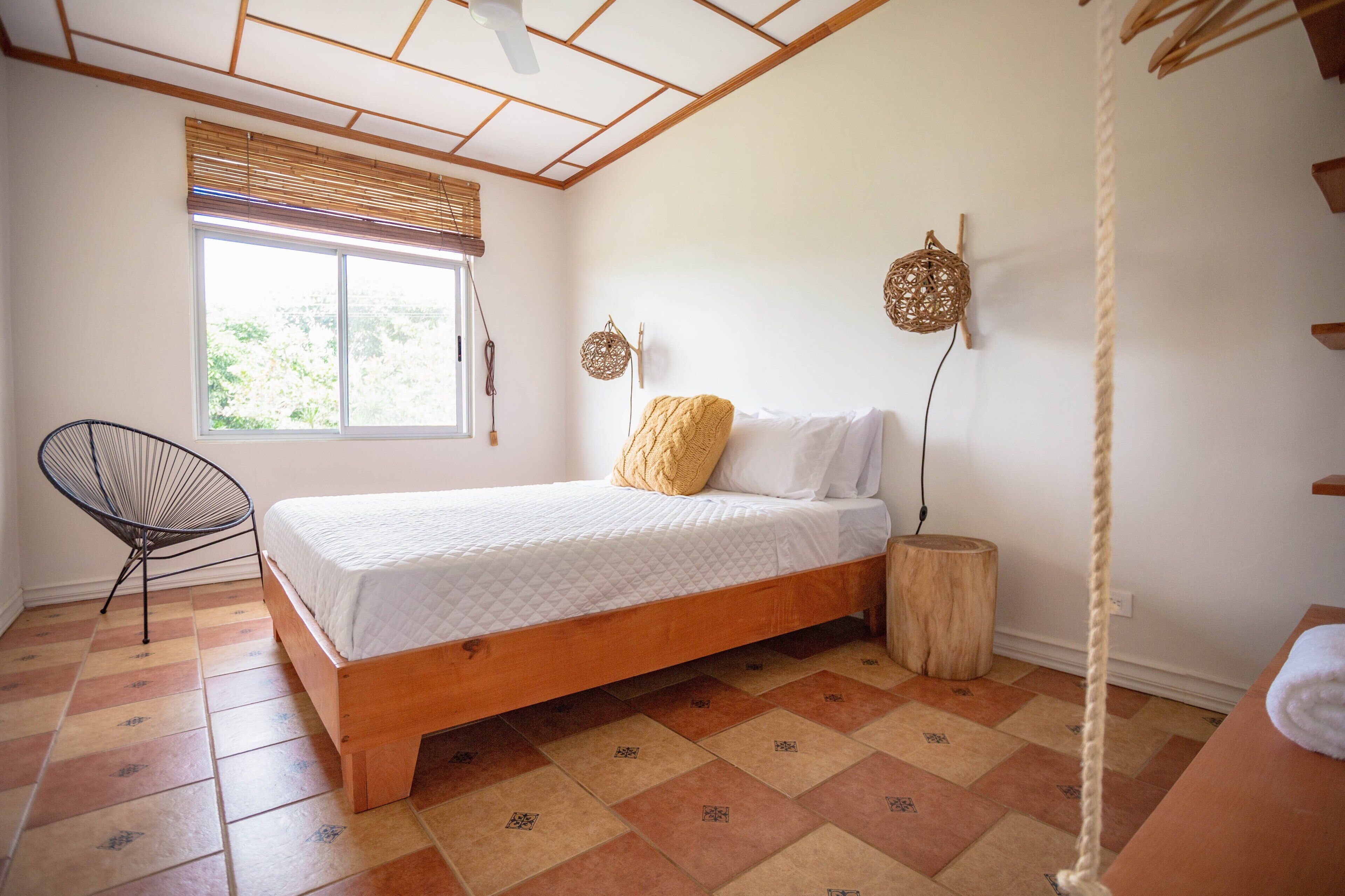 Ein Schlafzimmer mit einem hölzernen Bett, einem Sessel, einem Fenster mit Jalousien und einer daran befestigten Schnur.