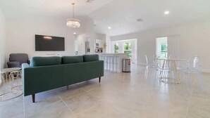 A spacious living area with a green sofa, a TV, and a dining area with clear chairs.