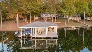 Una casa flotante amarrada en un lago, con terraza, asientos exteriores y un cobertizo pequeño.