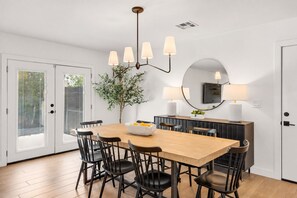 A dining area with a wooden table, black chairs, a bowl of fruit, a potted plant, and a modern chandelier.