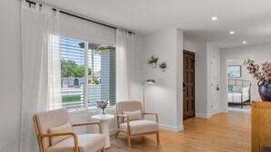 Ein modernes Wohnzimmer mit zwei beigefarbenen Sesseln, einem kleinen runden Tisch mit einer Vase und Blick auf einen Garten durch große Fenster mit weißen Vorhängen.