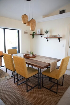 Un comedor con una mesa de madera, cuatro sillas, un mantel tejido, lámparas de mimbre colgantes y dos estantes empotrados con objetos decorativos.