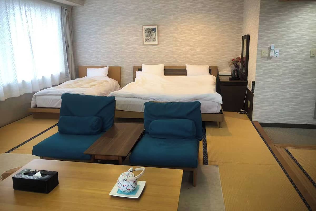 Hotelkamer met twee bedden, een klein tafeltje, twee stoelen, een nachtkastje met een vaas en een raam met gordijnen.