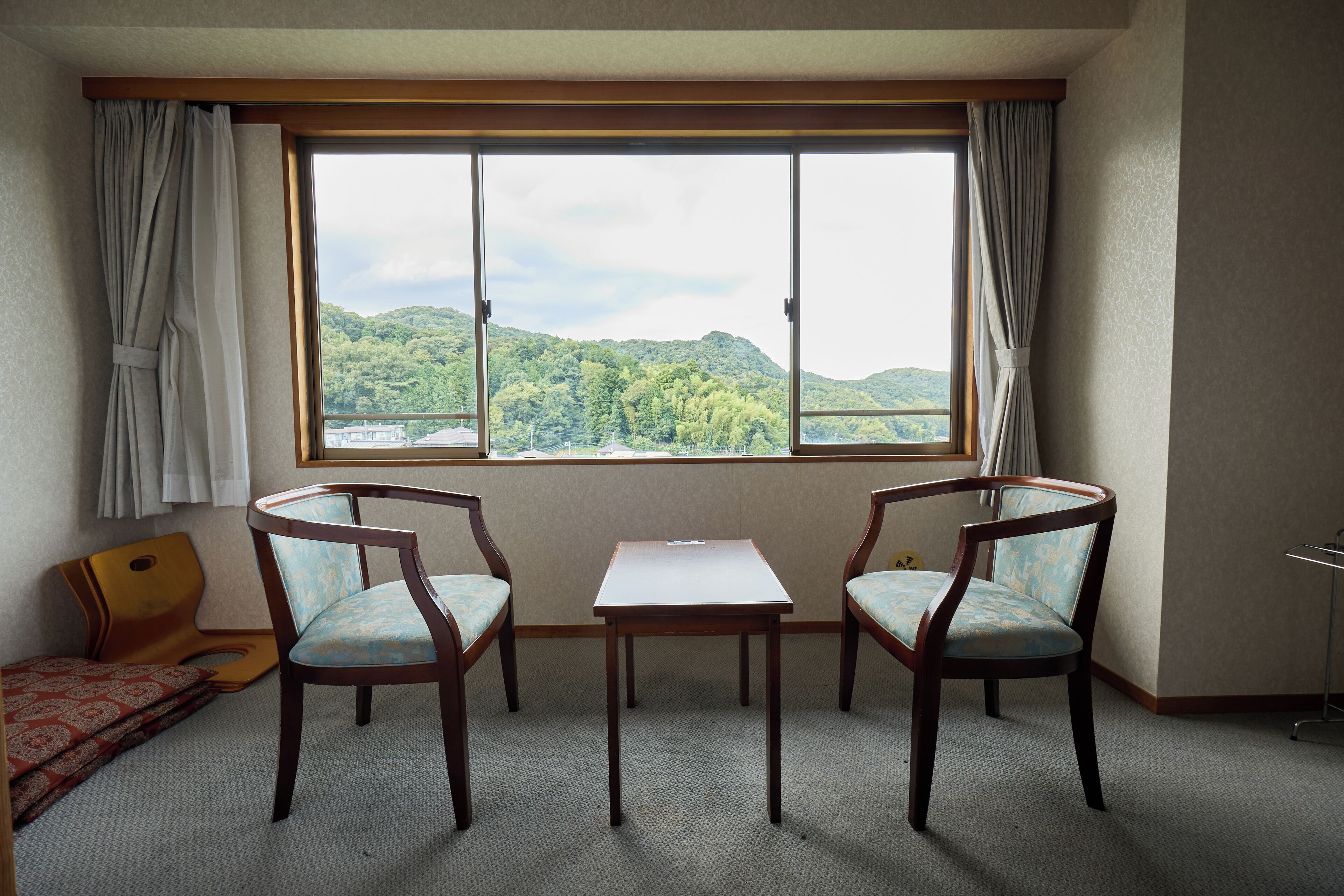 Une pièce dotée d’une grande fenêtre, de deux chaises en bois, d’une petite table et d’un lit recouvert d’une couvre-lit à motifs.