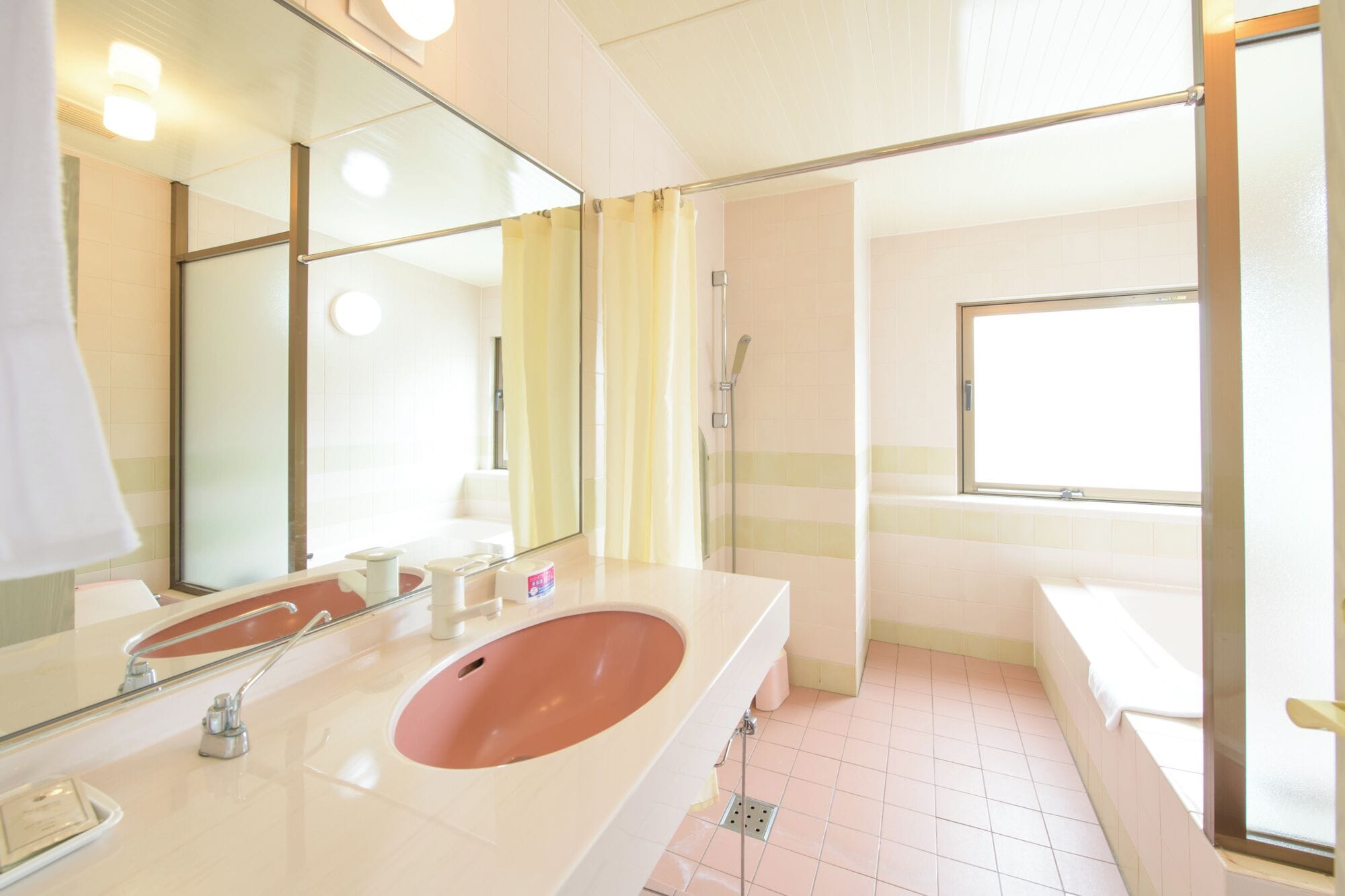 Un baño con un espejo grande, un lavabo con lavamanos rosa, zona de ducha y una ventana que proporciona luz natural.