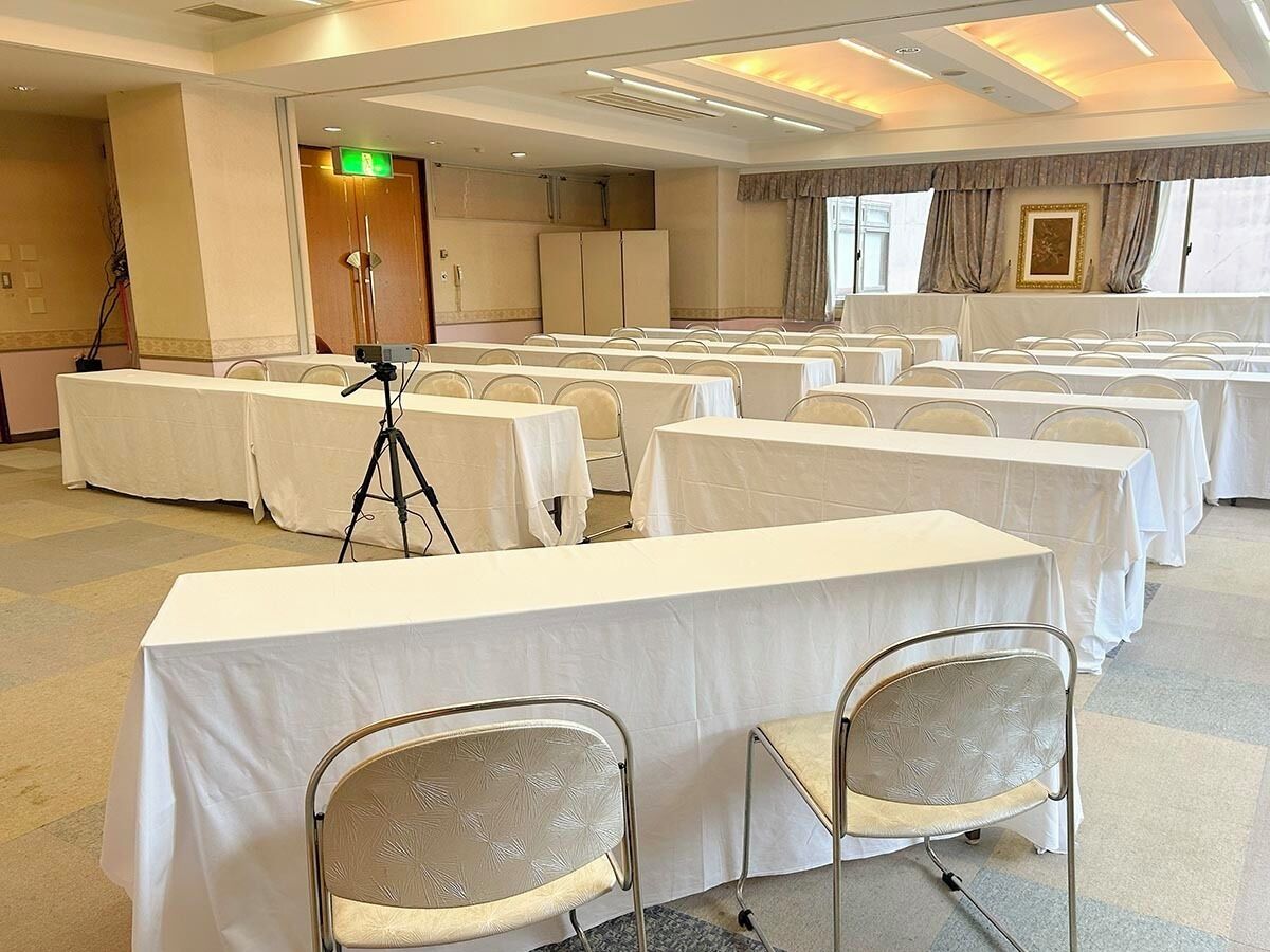 A conference room with multiple tables and chairs, a tripod, and a door with an emergency exit sign.