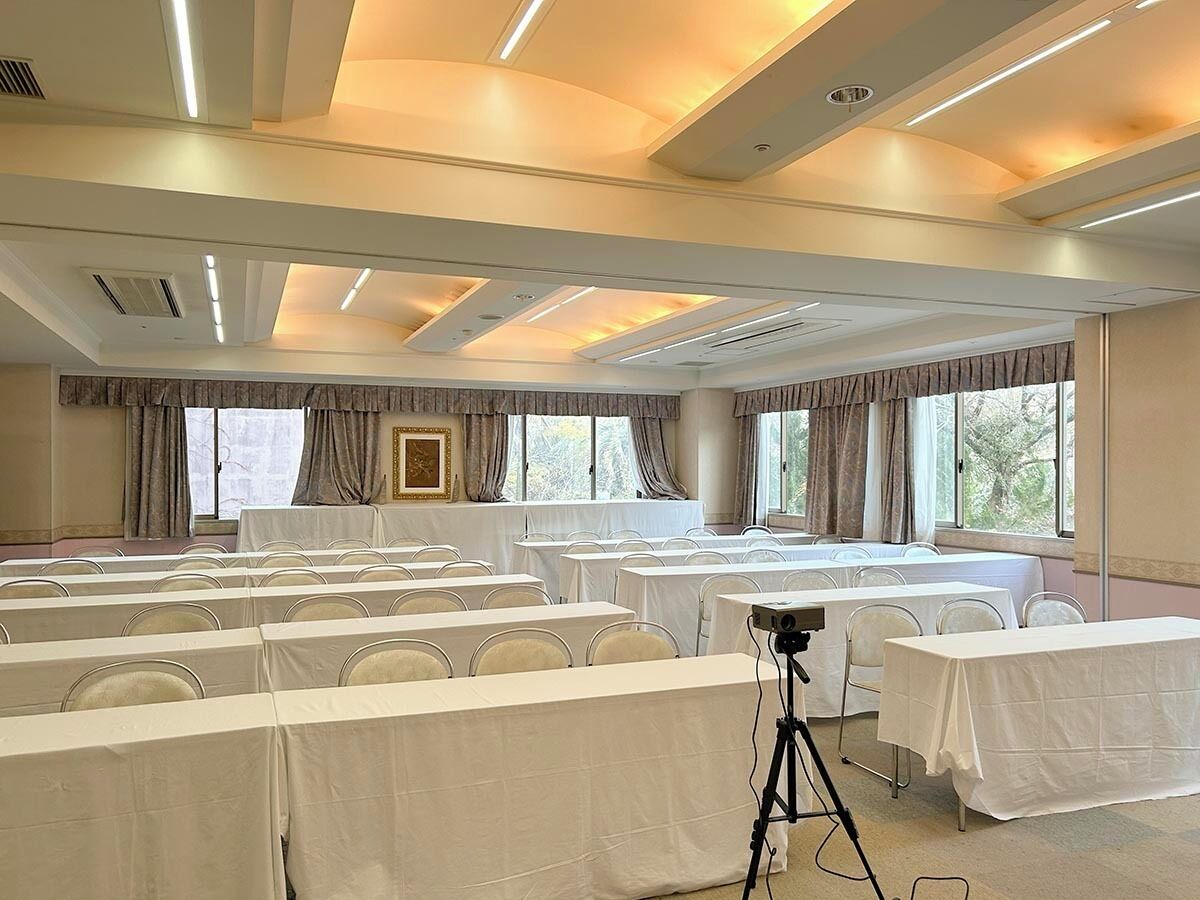 A conference room with rows of tables and chairs, large windows, and a ceiling with recessed lighting.