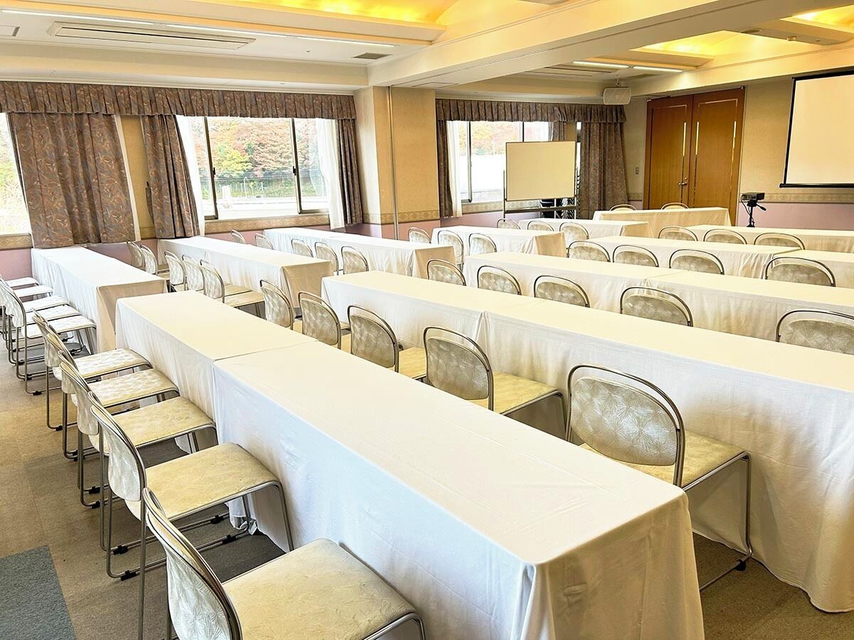 Une salle de conférence avec de longues tables et des chaises, de grandes fenêtres, des rideaux et un écran de projection.