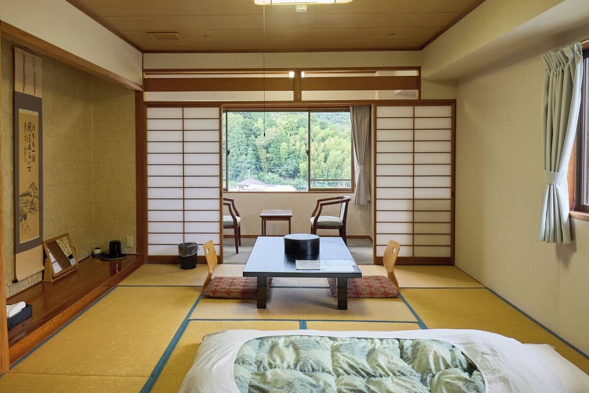 Een traditionele Japanse kamer met tatami vloerbedekking, een salontafel met kussens, een bed en uitzicht op een bos door een shoji scherm.