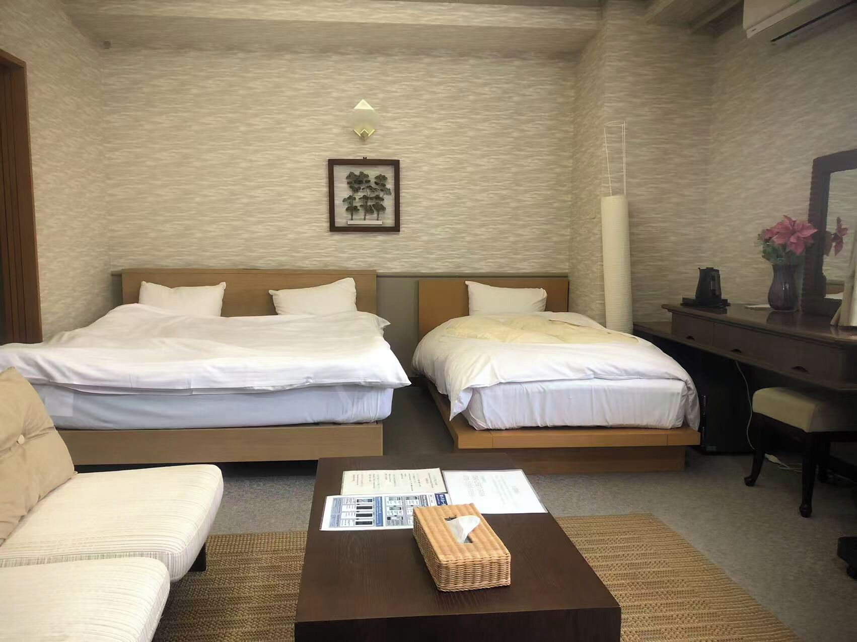 A hotel room with two beds, a desk, a chair, a coffee table with a tissue box, and a framed picture on the wall.