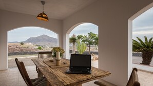 Una mesa de madera con una computadora portátil, rodeada de sillas, en una habitación con ventanales arqueados que dan a un paisaje con montañas y cactus.